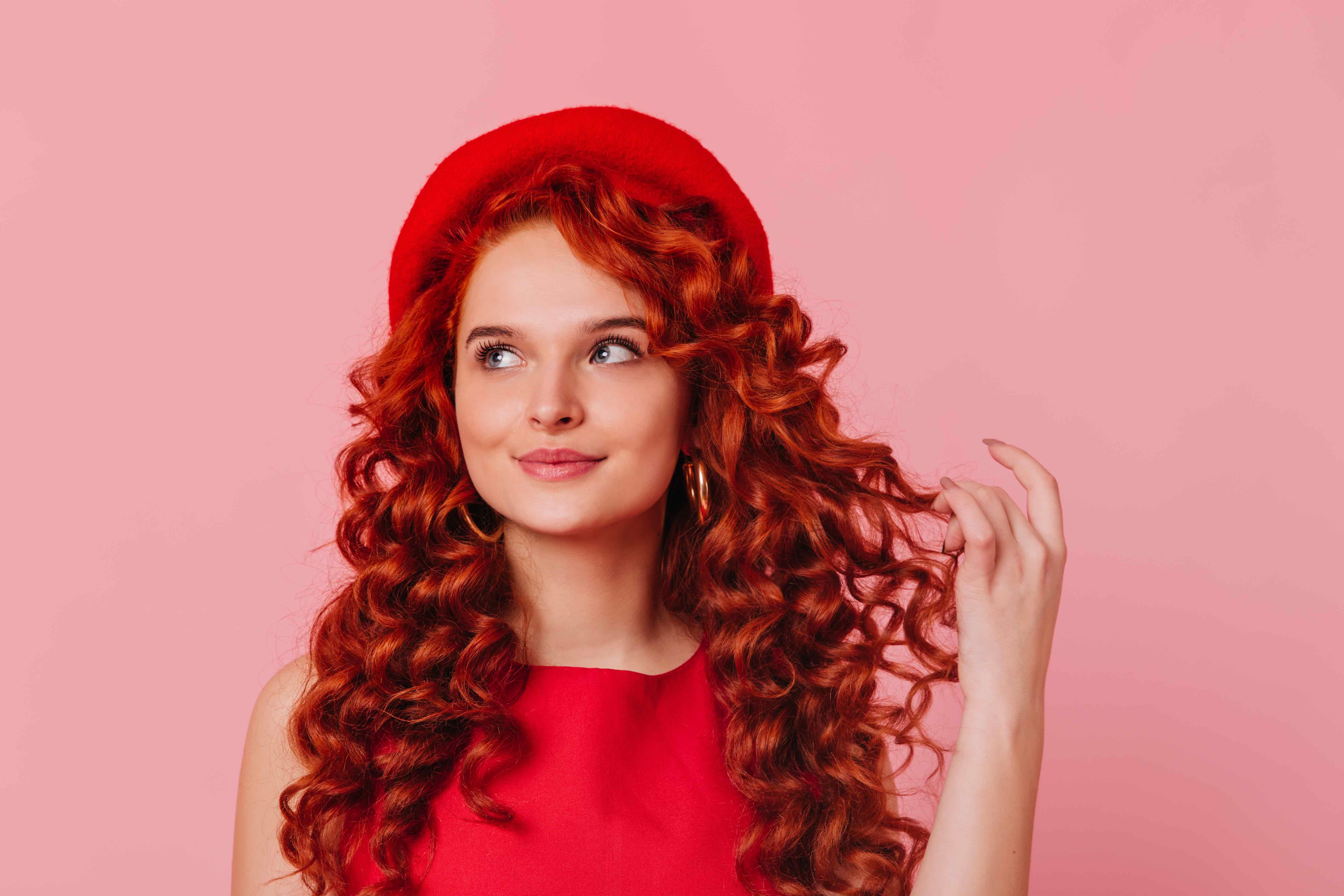 Close-up portrait of young stylish woman in red beret and top. Blue-eyed girl dreamily touches her red curls on pink background. Close-up portrait of young stylish woman in red beret and top. Blue-eyed girl dreamily touches her red curls on pink background